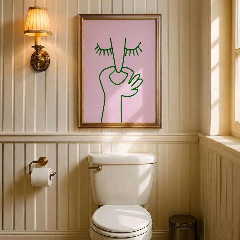 May include: A framed art print featuring a minimalist line drawing of a face with closed eyes and a hand, in green on a pink background. The artwork is in a gold frame, hanging above a white toilet in a bathroom.