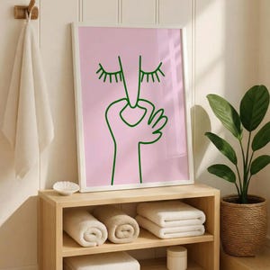 May include: A framed art print with a minimalist design. The artwork features a green line drawing of a face with closed eyes and a hand making an 'okay' gesture, set against a pink background. The print is displayed above a wooden shelf with rolled and folded towels.