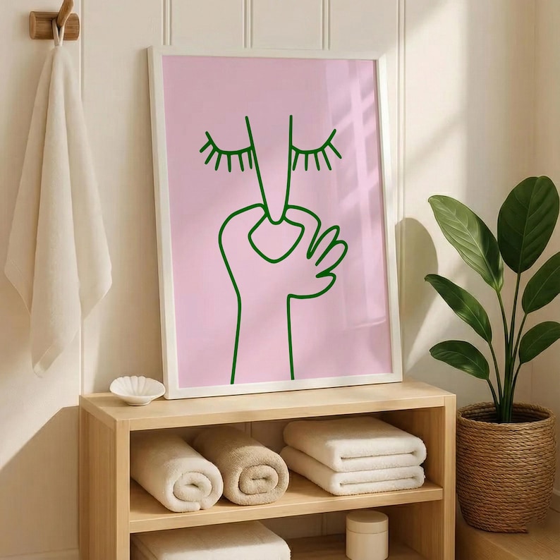 May include: A framed art print with a minimalist design. The artwork features a green line drawing of a face with closed eyes and a hand making an 'okay' gesture, set against a pink background. The print is displayed above a wooden shelf with rolled and folded towels.