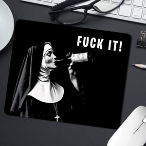 Puede incluir: Alfombrilla de ratón en blanco y negro con una monja bebiendo de una botella de vino. La frase "FUCK IT!" está impresa en blanco y negrita. La alfombrilla es rectangular y está sobre una superficie negra.