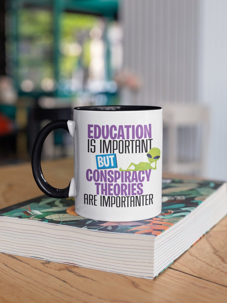 May include: White ceramic mug with a black rim and handle. The mug features a graphic of an alien with the text "Education is important but conspiracy theories are importanter".