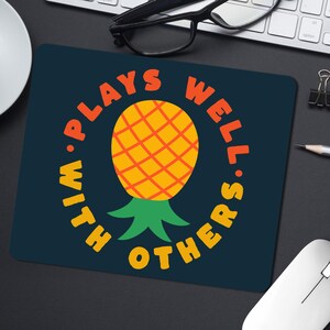May include: A rectangular mousepad with a dark blue background. The mousepad features a yellow and orange pineapple illustration in the center, surrounded by the words "PLAYS WELL WITH OTHERS" in orange. A white mouse and keyboard are visible.