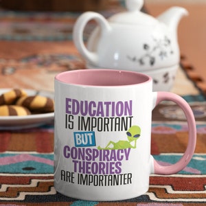 May include: White ceramic mug with a pink rim and handle. The mug features a graphic of a green alien with the text "Education is important but conspiracy theories are importanter".