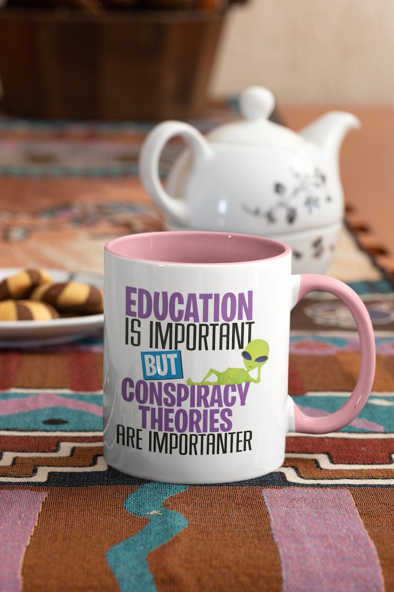 May include: White ceramic mug with a pink rim and handle. The mug features a graphic of a green alien with the text "Education is important but conspiracy theories are importanter".