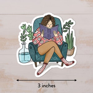 Puede incluir: Una pegatina con una ilustraci&oacute;n de dibujos animados de una persona sentada en un sill&oacute;n y leyendo un libro. La persona tiene el pelo casta&ntilde;o y lleva una camisa rosa con flores blancas. El sill&oacute;n es azul verdoso y hay dos plantas en macetas en el fondo. La pegatina mide 3 pulgadas de ancho.