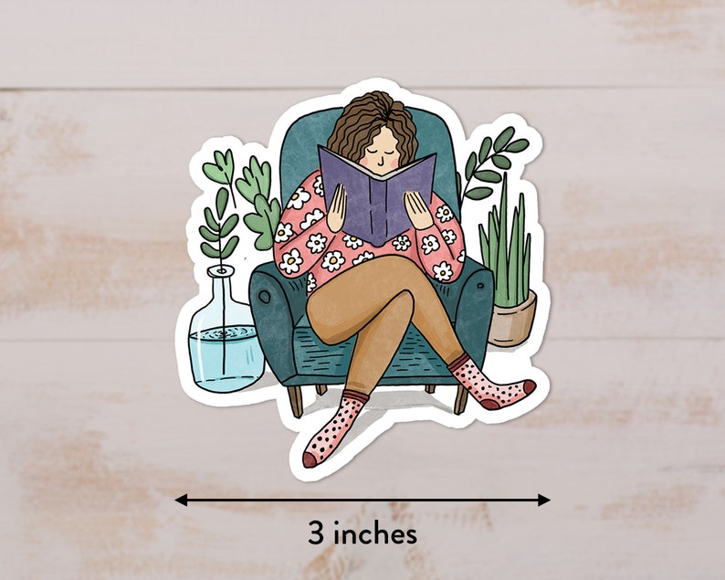 Puede incluir: Una pegatina con una ilustraci&oacute;n de dibujos animados de una persona sentada en un sill&oacute;n y leyendo un libro. La persona tiene el pelo casta&ntilde;o y lleva una camisa rosa con flores blancas. El sill&oacute;n es azul verdoso y hay dos plantas en macetas en el fondo. La pegatina mide 3 pulgadas de ancho.