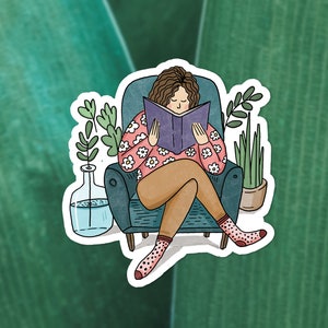 Puede incluir: Una mujer sentada en un sill&oacute;n azul lee un libro. Lleva un su&eacute;ter rosa con flores blancas y pantalones marrones. Hay dos plantas en macetas en la habitaci&oacute;n.