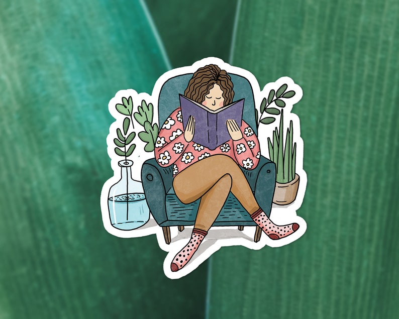 Puede incluir: Una mujer sentada en un sill&oacute;n azul lee un libro. Lleva un su&eacute;ter rosa con flores blancas y pantalones marrones. Hay dos plantas en macetas en la habitaci&oacute;n.