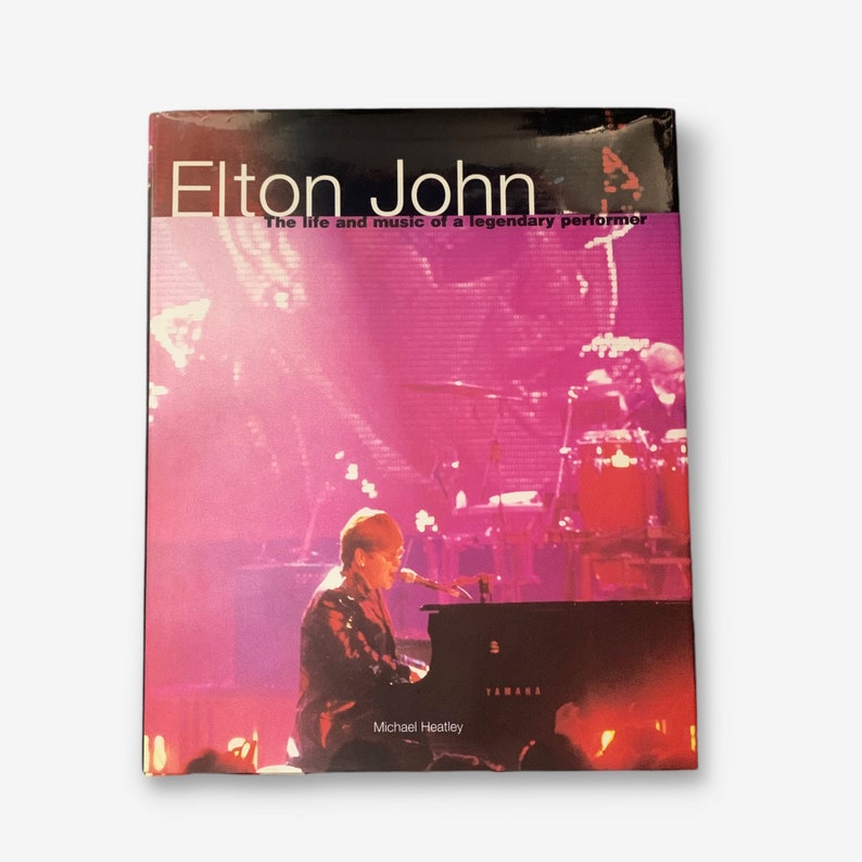 May include: A book cover featuring a photo of Elton John performing on stage. The cover is black with white text that reads "Elton John" and "The life and music of a legendary performer". The photo shows Elton John playing a black Yamaha piano with a microphone in front of him. The background is a pink and purple stage with a drummer and other musicians in the background.