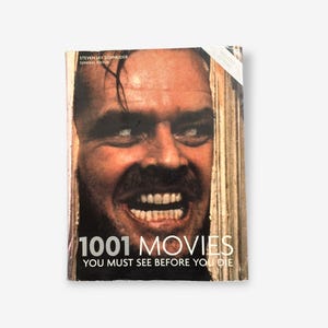 Può includere: Una copertina di libro con un primo piano del volto di Jack Nicholson dal film "Shining". Il titolo del libro è "1001 film da vedere prima di morire" e il testo "Edizione aggiornata" è nell'angolo in alto a destra.