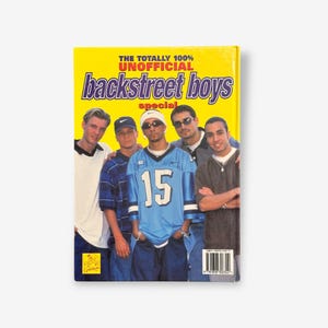 May include: A yellow book cover with the title "The Totally 100% Unofficial Backstreet Boys Special". The cover features a photo of five men, all wearing casual clothing. The man in the center is wearing a blue jersey with the number 15 on it.