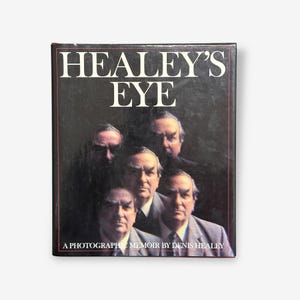 Pode incluir: Um livro com o título "HEALEY'S EYE" com uma capa preta. A capa apresenta um retrato de vários homens de fato. O texto "A PHOTOGRAPHIC MEMOIR BY DENIS HEALEY" está na parte inferior.