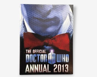 May include: The Official Doctor Who Annual 2013 book cover features a close-up of the Doctor's face with a red bow tie. The cover is black with white text.