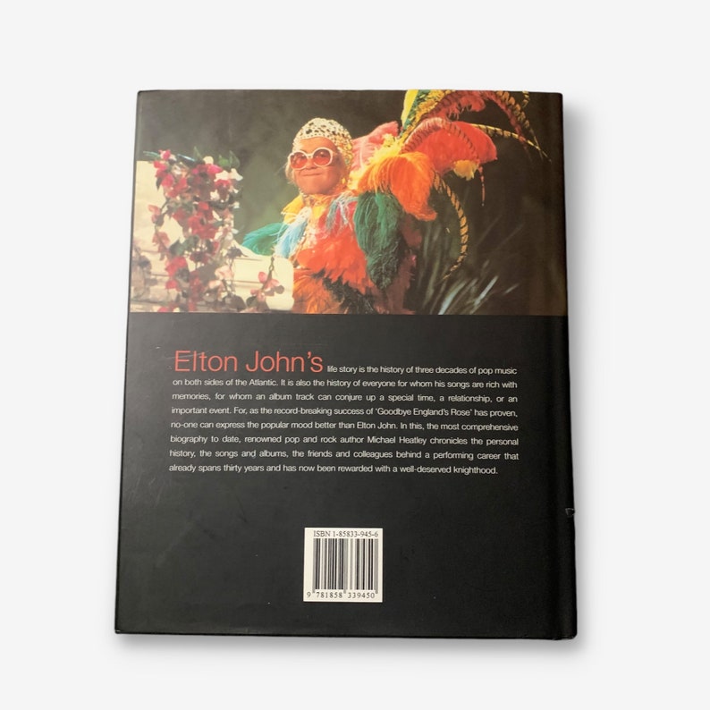 May include: A black and white photo of Elton John wearing a colorful feathered costume and sunglasses. The back cover of a book titled "Elton John's" with a description of the book. The ISBN number is 1-85833-945-6.