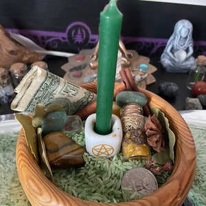 May include: A wooden bowl filled with green rice containing a green taper candle in a white pentacle candle holder, a rolled one-dollar bill, a small glass vial with herbs, a tiger eye stone, a green stone, a star anise, and a silver quarter.