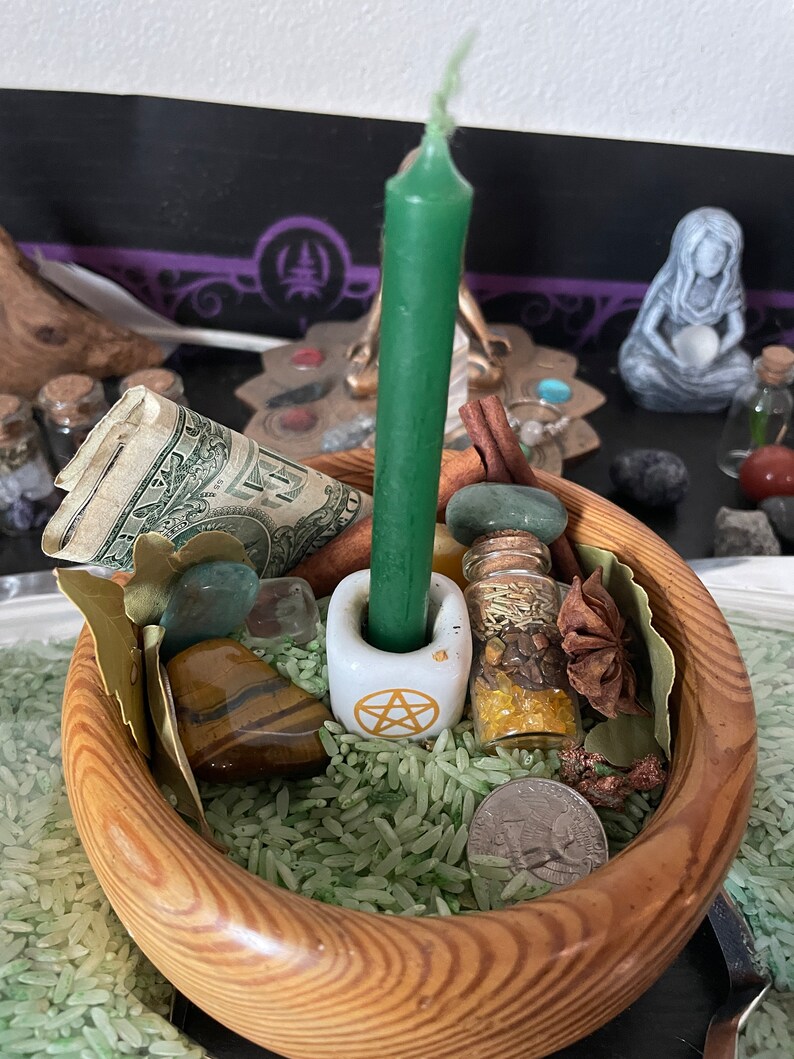 May include: A wooden bowl filled with green rice containing a green taper candle in a white pentacle candle holder, a rolled one-dollar bill, a small glass vial with herbs, a tiger eye stone, a green stone, a star anise, and a silver quarter.