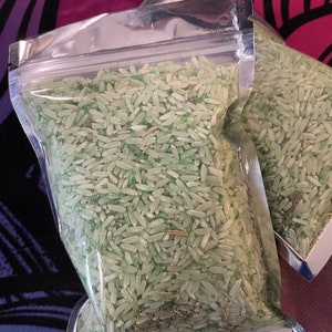 May include: Two clear, resealable plastic pouches filled with long-grain white rice that has been dyed a pale mint green color. The rice grains are mixed with small, thin, dried brown botanical pieces throughout the contents of the bags.