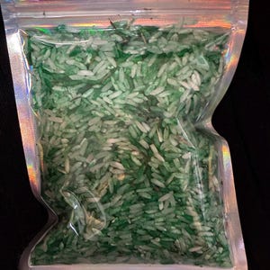 May include: A clear, holographic-edged resealable plastic pouch filled with a mixture of white and various shades of green dyed rice grains. The bag features a top hanging hole and a zip-lock closure, containing a loose collection of small, elongated, dried food-grade rice kernels.