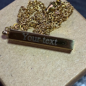 May include: A gold-toned metal bar pendant necklace featuring a delicate chain and a rectangular charm engraved with the words Your text. The piece includes a lobster claw clasp and is presented with the chain coiled behind the horizontal bar.