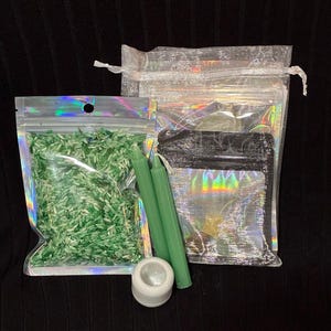 May include: A set containing a clear holographic pouch filled with green rice, two slender sage green taper candles, a small white cylindrical candle holder, and a larger translucent white drawstring bag with a black interior pouch.