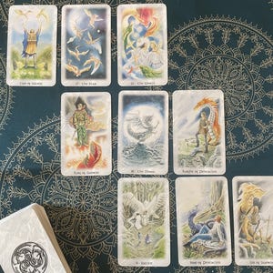 May include: A spread of tarot cards featuring illustrations of dragons, birds, and other mythical creatures. The cards are laid out in a grid pattern on a blue and gold patterned surface. The cards include "The Star", "The Moon", "Knight of Pentacles", "Nine of Pentacles", and "Six of Swords".