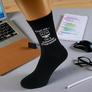 May include: Black socks with the text "Trust me... I'm a Computer Programmer" in white, featuring a computer monitor graphic. The socks are on a person's leg, with a laptop, mouse, and glasses in the background.