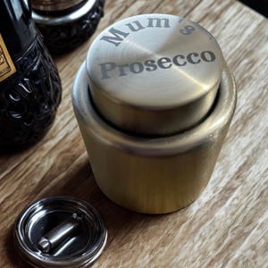 May include: A gold-toned Prosecco bottle stopper, engraved with "Mum's Prosecco". The stopper is cylindrical and sits on a wooden surface. A bottle of Prosecco is visible in the background, along with a detached stopper base.