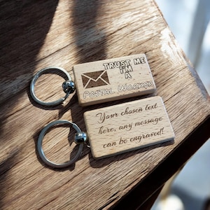 May include: Two wooden keyrings with silver key rings. The top keyring has a small envelope icon and the text "TRUST ME I'M A POSTAL WORKER". The bottom keyring has the text "Your chosen text here, any message can be engraved!"