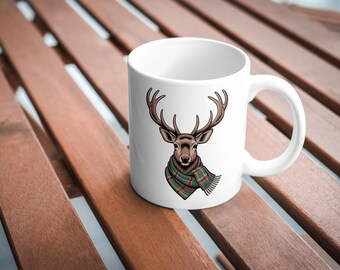 May include: White ceramic mug with a deer head illustration, complete with antlers and a green and red plaid scarf. The mug sits on a wooden surface. Ideal for hot beverages, such as tea or coffee.