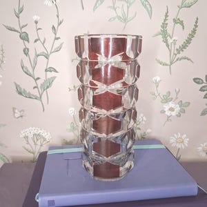 May include: A cylindrical glass vase featuring a repeating geometric pattern of raised, clear, hexagonal facets. The interior of the vase has a deep, dark red hue, creating a contrast with the transparent, textured exterior surface of the glass.