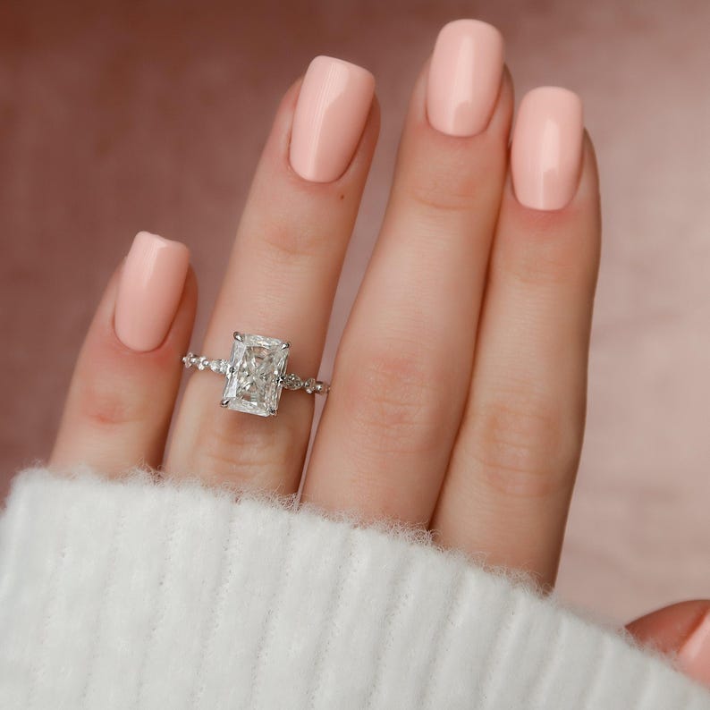 May include: A close-up of a hand wearing a silver ring with a large, rectangular, clear diamond. The hand is wearing light pink nail polish.