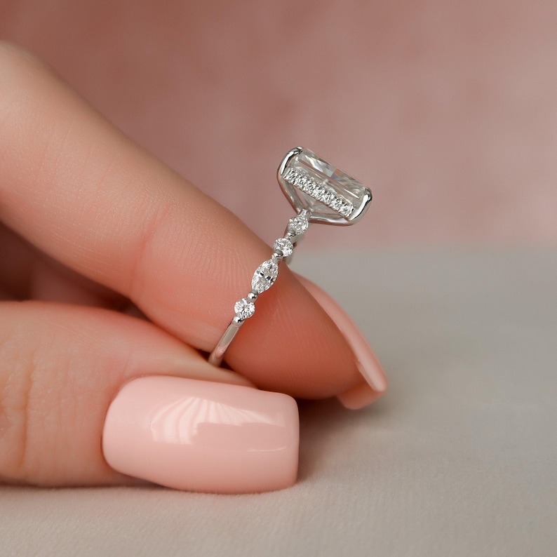 May include: A silver ring featuring a large, clear, rectangular-shaped gemstone. The gemstone is surrounded by smaller, clear gemstones. The band of the ring is set with smaller, clear gemstones.