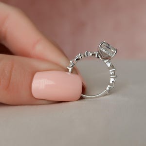 May include: A silver ring featuring a large, clear, square-shaped gemstone surrounded by smaller diamonds. The ring has a delicate, intricate design.