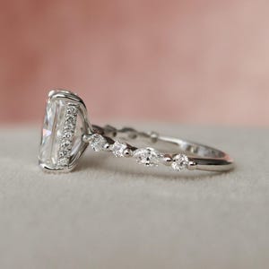May include: A silver engagement ring featuring a large, rectangular, clear gemstone surrounded by smaller clear gemstones. The band is decorated with smaller, pear-shaped clear gemstones.