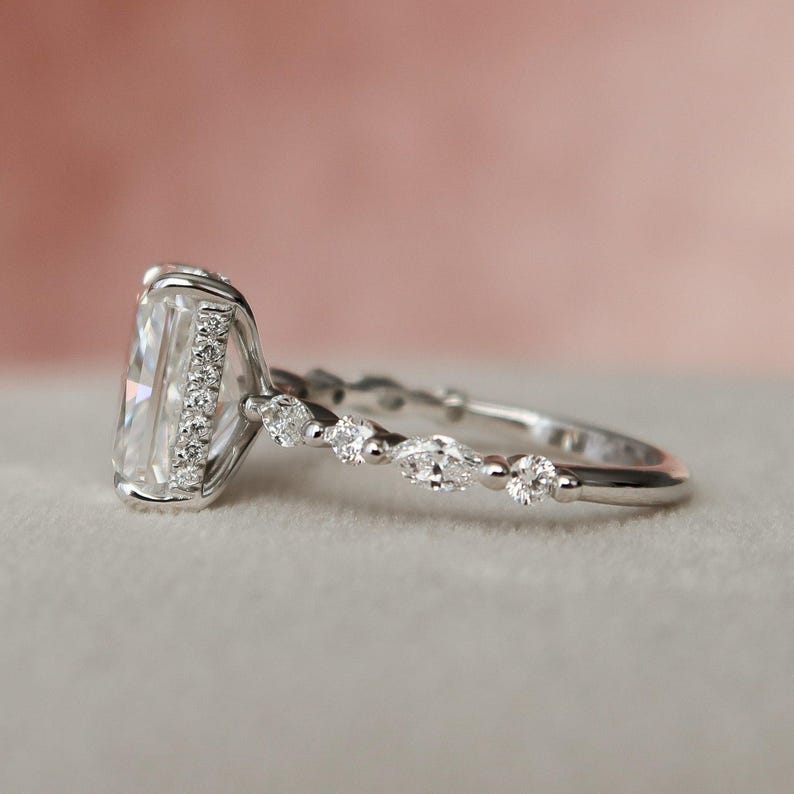 May include: A silver engagement ring featuring a large, rectangular, clear gemstone surrounded by smaller clear gemstones. The band is decorated with smaller, pear-shaped clear gemstones.