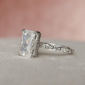 May include: A silver engagement ring featuring a large, rectangular, clear gemstone surrounded by smaller gemstones. The band is adorned with a row of smaller, oval-shaped gemstones.