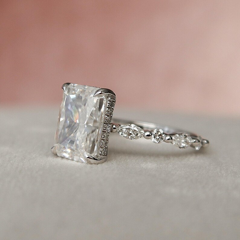 May include: A silver engagement ring featuring a large, rectangular, clear gemstone surrounded by smaller gemstones. The band is adorned with a row of smaller, oval-shaped gemstones.