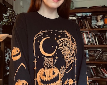 May include: Black long-sleeve shirt with a Halloween-themed design. The orange graphic features a cat in a pumpkin carriage, a spider web, a crescent moon, and stars. The sleeves have pumpkin and scythe designs.