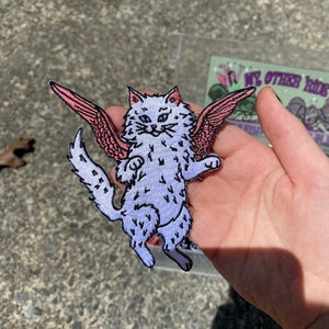 May include: Embroidered patch of a light purple cat with pink and red wings. The cat has blue eyes and a fluffy tail. The patch is held in a hand against a gray background. The text "MY OTHER RIDE" is visible on a card.