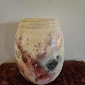 Puede incluir: Un jarrón de cerámica redondeado con una base de color crema que presenta manchas moteadas de rojo oscuro, negro y verde apagado. La parte superior del jarrón tiene una serie de estrías horizontales que rodean el cuello de la pieza.