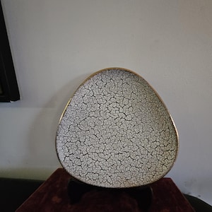 May include: A triangular-shaped decorative plate featuring a white surface covered in a dense, black crackle pattern. The plate has a smooth, metallic-toned rim that follows its rounded edges, creating a distinct contrast against the textured finish.