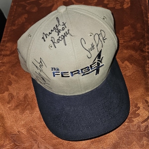 May include: A tan baseball cap with a dark blue brim featuring the embroidered text FAB FERBEY 4 in blue and white. The crown of the cap is covered in multiple black ink signatures, including names like Marcel Rocque and Scott Pfeifer written clearly.