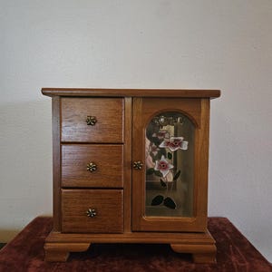 Peut inclure: Une armoire à bijoux en bois dotée de trois tiroirs superposés sur la gauche et d'une porte sur la droite. La porte comporte un panneau en verre orné d'un motif floral peint en blanc et rose. Chaque tiroir et la porte possèdent des boutons en forme de fleur.