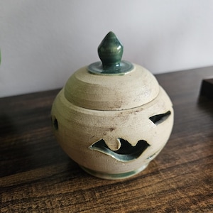 May include: A round, beige ceramic jar with a removable lid featuring a pointed green handle. The body of the jar has carved, open cutouts in a wave-like pattern around the centre, revealing a dark interior. The base also shows green glaze.