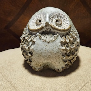 May include: A round, ceramic owl figurine with a speckled light gray and brown finish. The owl features deeply carved, textured feathers on its sides, prominent circular eyes, and a distinct brow line, all rendered in a matte, earthy glaze.