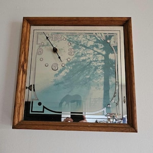 May include: A square wall clock with a wooden frame and a mirrored glass face. The clock features a printed image of a tree and a horse in a field. The dial includes black numbers at the 3, 6, 9, and 12 positions, with black hands indicating the time.