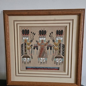 Peut inclure: Une œuvre d'art en sable encadrée représentant trois personnages stylisés avec des coiffes à plumes et des motifs géométriques dans des tons de brun, noir, blanc et bleu. L'œuvre est insérée dans un cadre en bois brun clair avec un passe-partout beige.
