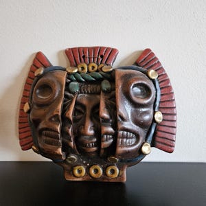 May include: A brown ceramic wall mask featuring three distinct faces layered together. The outer faces are flanked by red, fan-shaped side panels. The central face is framed by a green braided element, and the entire piece is accented with small, circular gold-colored studs along the edges and base.