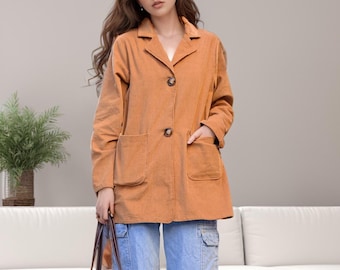Könnte beinhalten: Eine Frau trägt einen orangefarbenen Cord-Blazer mit zwei großen Vordertaschen und zwei Knöpfen. Sie trägt hellblaue Cargo-Jeans und weiße Turnschuhe. Sie hält eine schwarz-weiß karierte Tragetasche mit braunen Griffen.