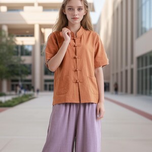 Pode incluir: Uma mulher vestindo uma camisa de manga curta de cor laranja claro com colarinho mandarim e uma fileira de bot&otilde;es na frente. Ela tamb&eacute;m est&aacute; vestindo cal&ccedil;as largas de cor lavanda e sapatos brancos.
